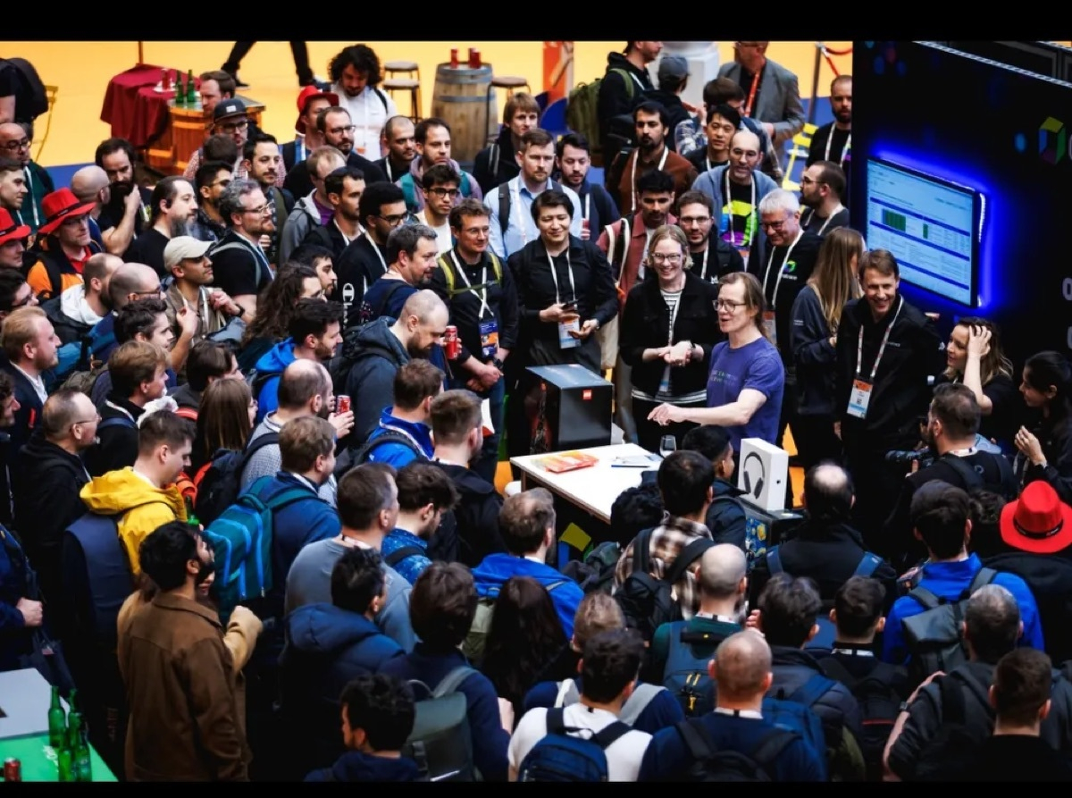 Crowd of conference attendees packed shoulder-to-shoulder around a booth at a tech event, with a presenter in a blue shirt at the center speaking to the audience